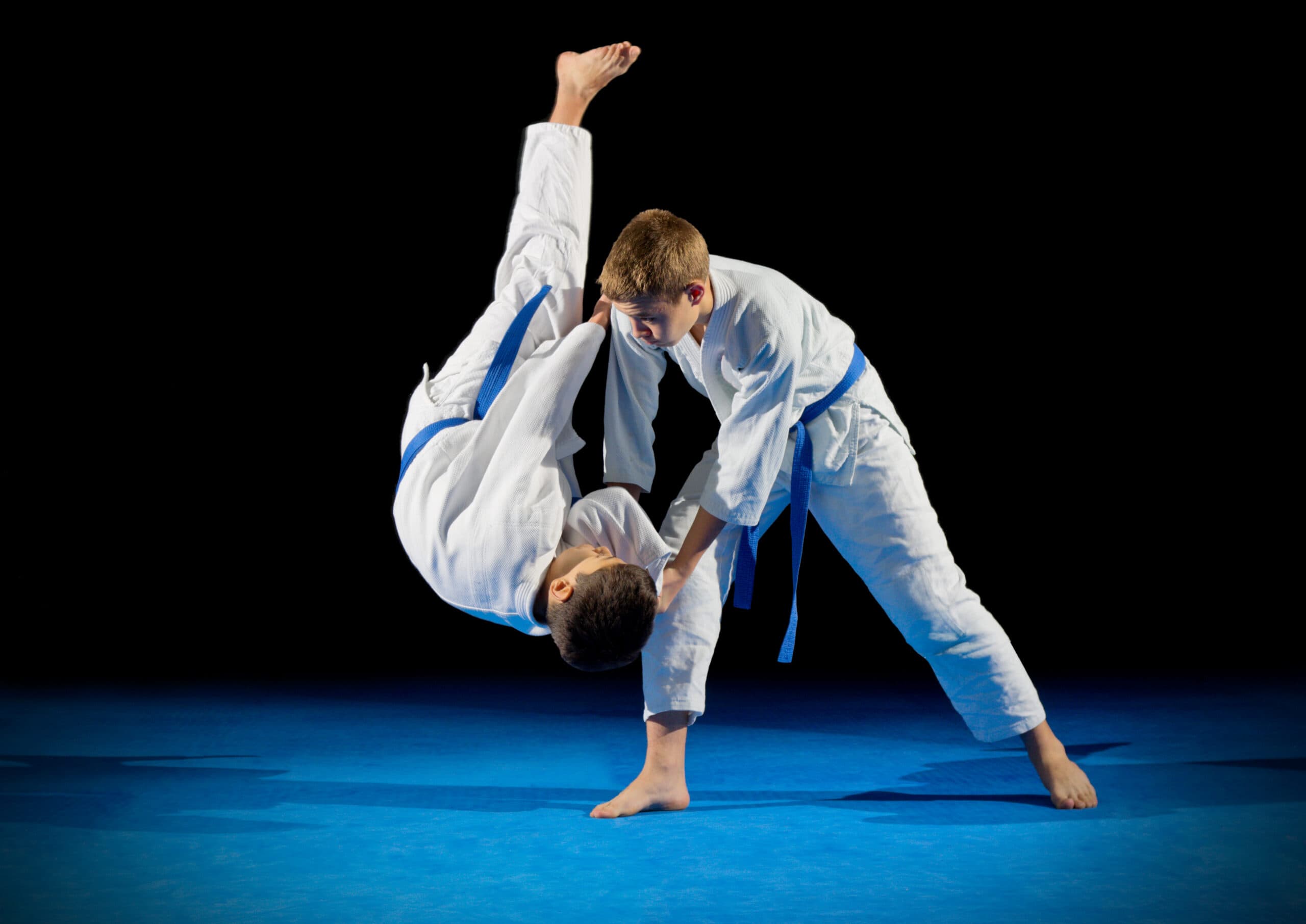 Boy performing a martial arts throw on another student during a judo training session, both wearing white uniforms with blue belts.